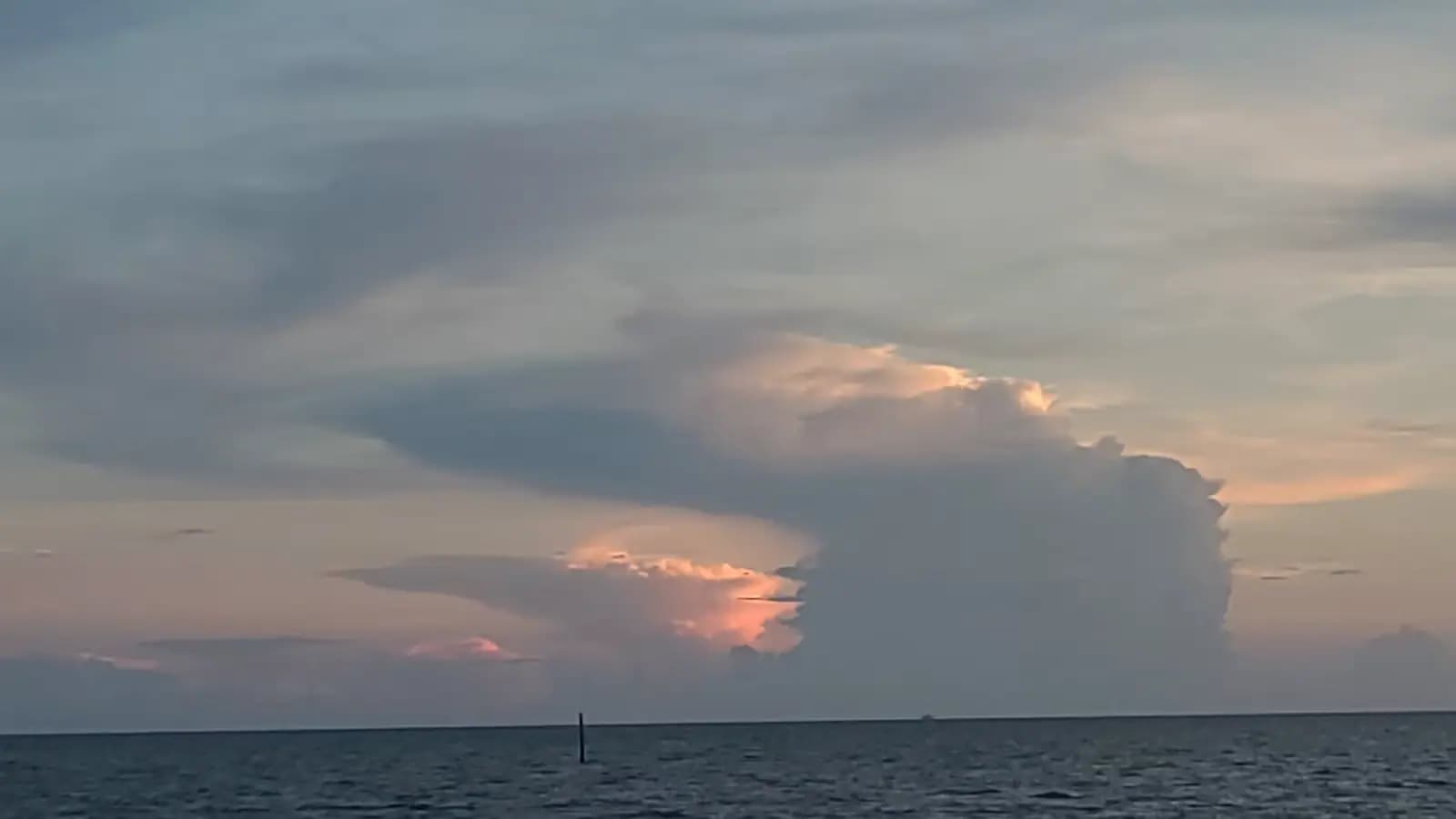 A tall thunderhead building over flat coastal water at dusk with a single channel marker in the foreground