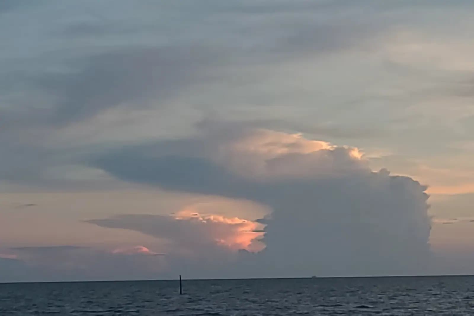 A tall thunderhead building over flat coastal water at dusk with a single channel marker in the foreground