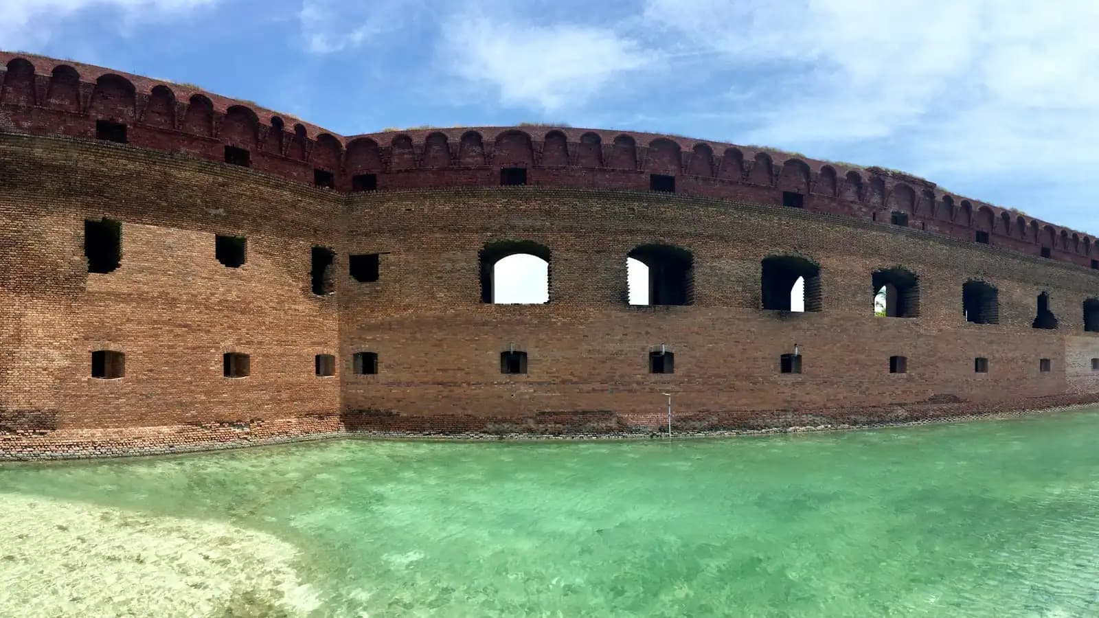 A red-brick coastal fort wall rising straight out of shallow green water under a blue sky