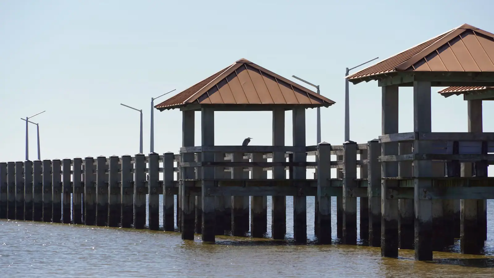 A long low fishing pier with covered shelters standing over flat brown water under a pale sky