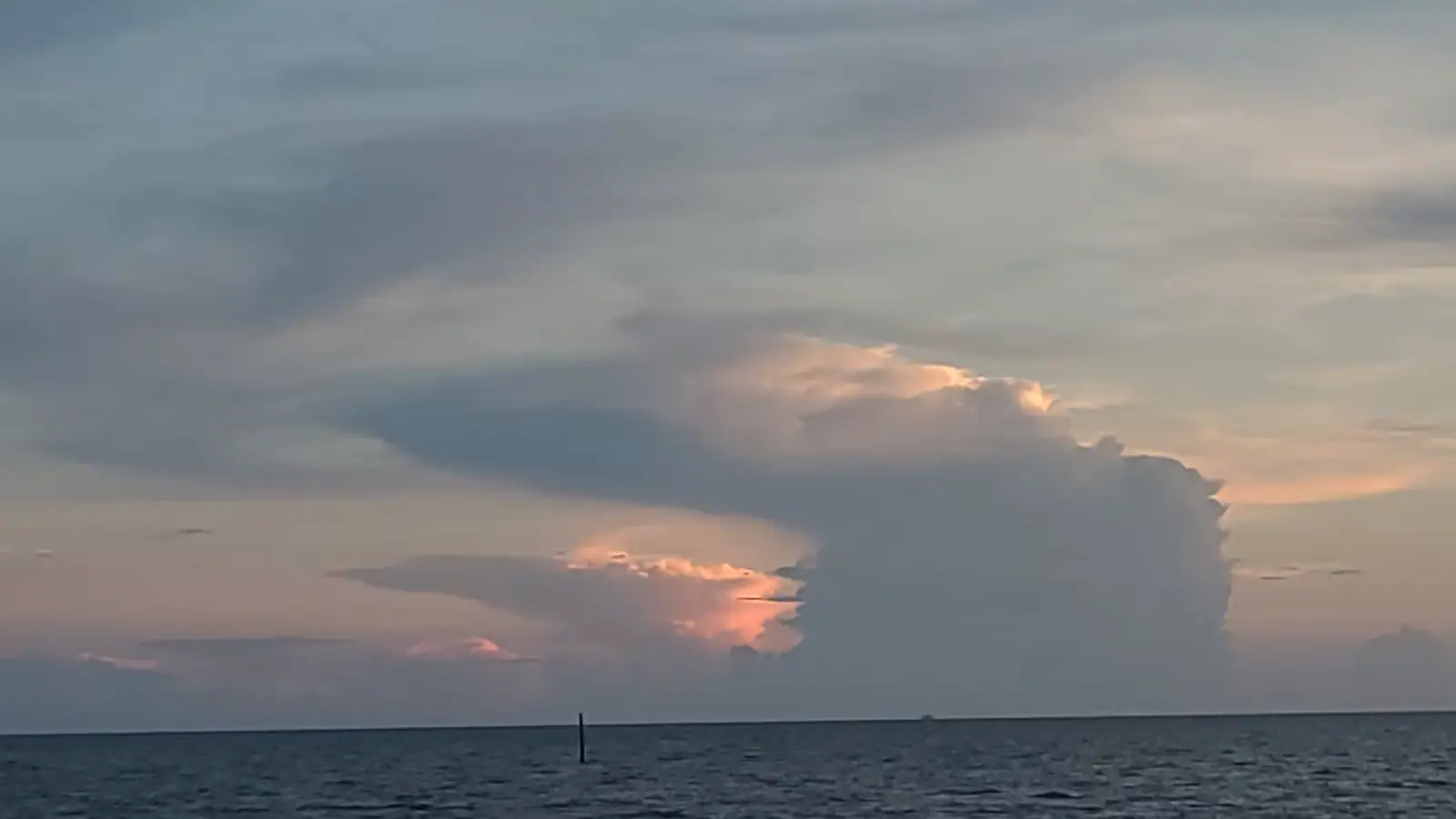 A tall thunderhead building over flat coastal water at dusk with a single channel marker in the foreground