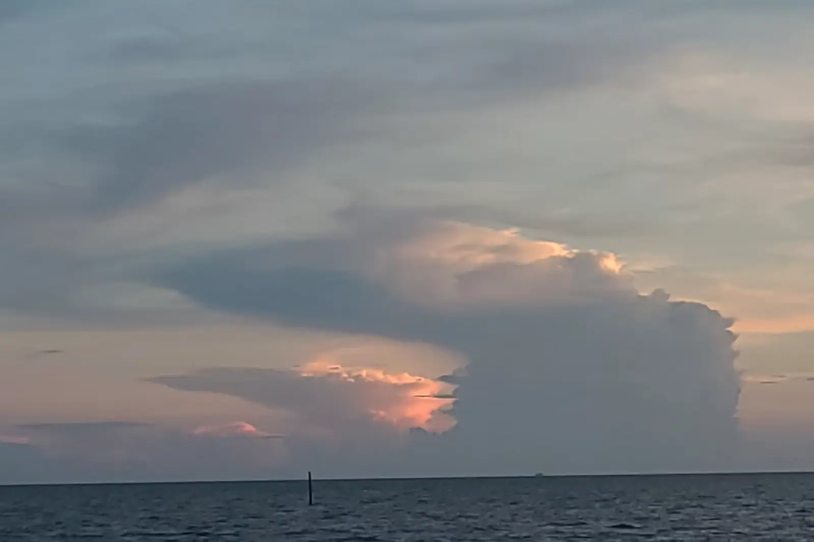 A tall thunderhead building over flat coastal water at dusk with a single channel marker in the foreground