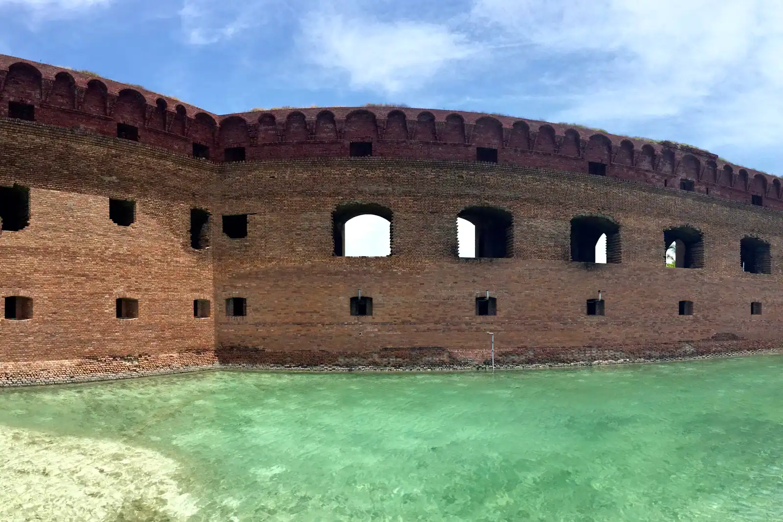 A red-brick coastal fort wall rising straight out of shallow green water under a blue sky